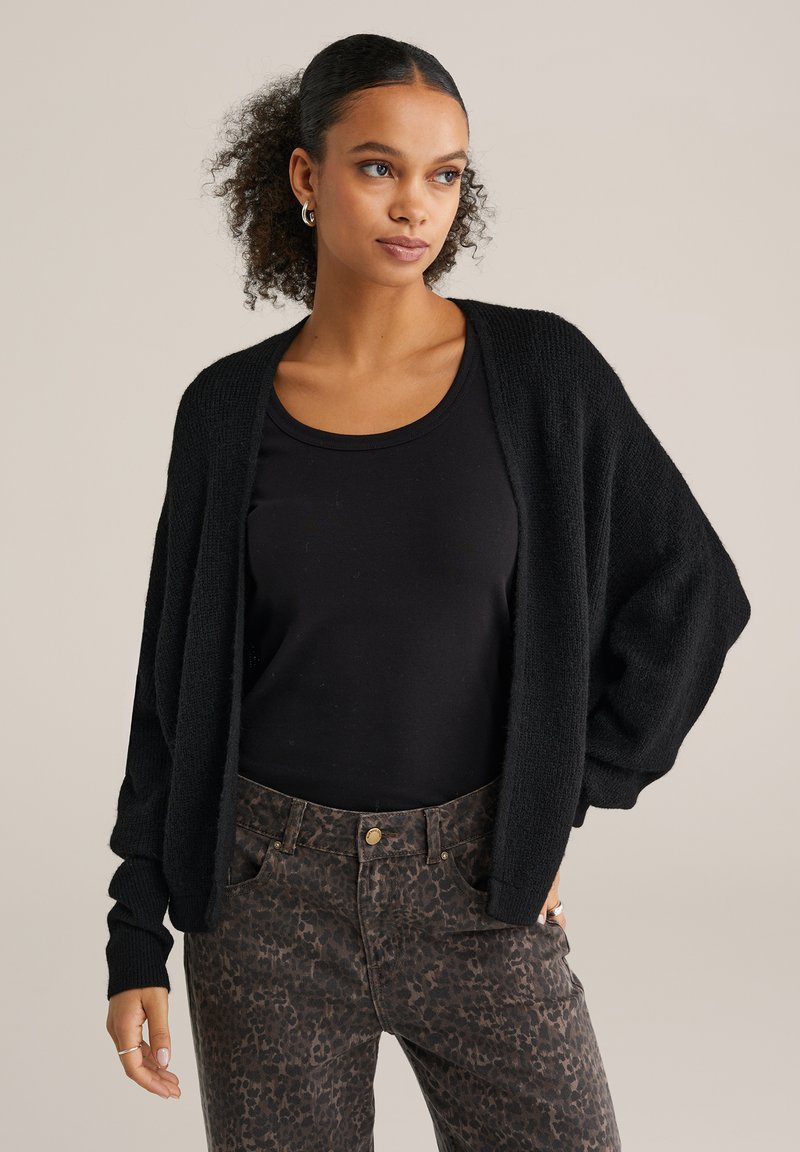 Cardigan nero in maglia con vestibilità morbida e maniche larghe, abbinato a una maglietta nera e pantaloni marroni con stampa maculata. Texture liscia.