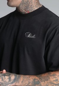 Camiseta de algodón negra con un ajuste relajado, que presenta un logo en cursiva blanca en el lado izquierdo del pecho y mangas cortas. Textura suave.