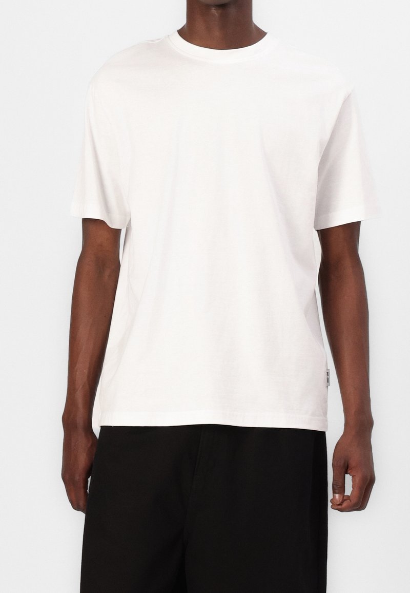 Homme portant un T-shirt blanc uni à manches courtes et col rond, ainsi qu'un pantalon noir, debout devant un fond blanc.