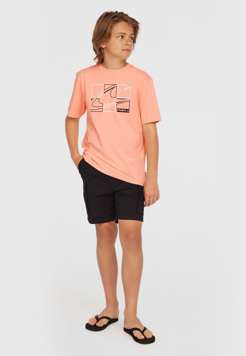 Camiseta corta de manga en color naranja claro con un diseño gráfico negro, combinada con pantalones cortos de cargo negros y chanclas negras. Atuendo casual de verano.