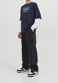 Marinblå grafisk t-shirt över en vit långärmad topp, kombinerad med lösa svarta jeans och svart-vita sneakers.