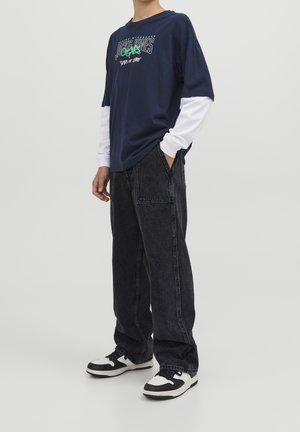 T-shirt grafica blu marina sovrapposta a una maglietta bianca a maniche lunghe, abbinata a jeans neri larghi e sneakers bianco e nere.