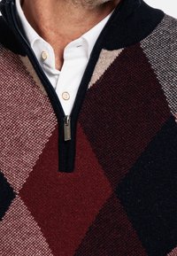 Sweter z zamkiem błyskawicznym, sięgający do ćwierci, w plecionym wzorze diamentów; kolory obejmują burgund, granat, beż i krem. Posiada teksturowany materiał i kołnierz.