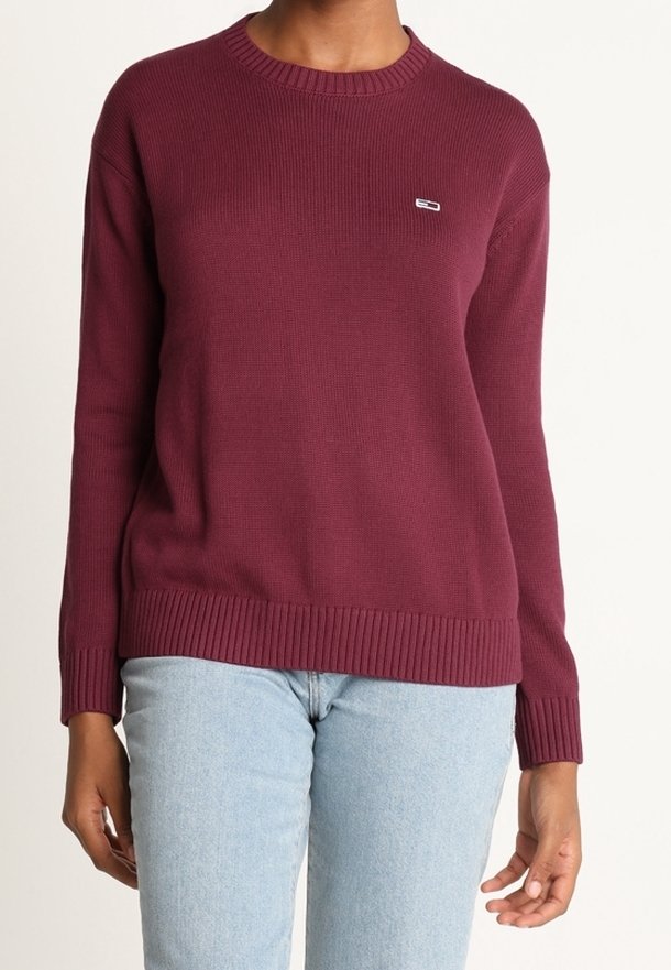 Personne portant un pull en maille bordeaux uni et un jean bleu clair, debout devant un fond neutre.