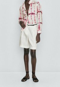 Short-sleeve shirt in cream with pink tie-dye pattern, paired with white linen shorts. Black belt and strappy black sandals complete the look.