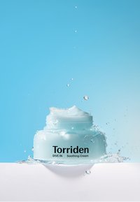 Valkoinen vaaleansininen purkki Torriden Dive In Soothing Creamia, jossa on sileä koostumus, mustatekstinen etiketöinti ja vesipisaroita sinistä taustaa vasten.