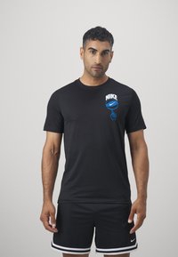 Schwarzes Baumwoll-T-Shirt mit Rundhalsausschnitt, mit einem weißen und blauen Nike-Logo und Grafik auf der linken Brust. Das Modell trägt schwarze Shorts mit weißen Akzenten.