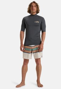 Grå kortärmad surftröja med hög krage, tillsammans med krämfärgade badshorts med horisontella ränder i grön, orange och blå.