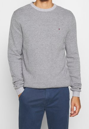 Mann trägt einen hellgrauen strukturierten Rundhalspullover mit kleinem Logo auf der Brust und marineblaue Hose, Hände entspannt an den Seiten.