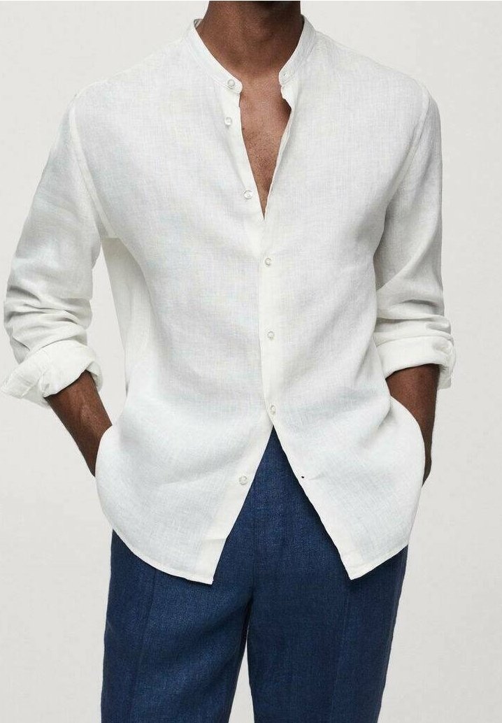 Homme portant une chemise blanche à manches longues boutonnée avec les manches retroussées et un pantalon bleu, les mains dans les poches, sur un fond uni.