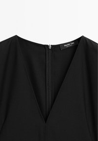 Svart klänning med djup V-ringning och dragkedja bak; slät, texturerad tyg ger en åtsittande silhuett och rena linjer; varumärkesetikett synlig.