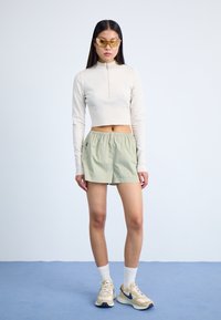 Cream-colored, fitted long-sleeve top with zipper; light green, elastic-waist shorts; yellow sunglasses; beige sneakers with navy accents.