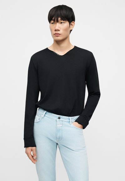 Νεαρός άνδρας που φοράει μαύρο μακρυμάνικο πουκάμισο με μικρά κουμπιά στο ντεκολτέ και ανοιχτό μπλε τζιν, στέκεται με το ένα χέρι στην τσέπη.