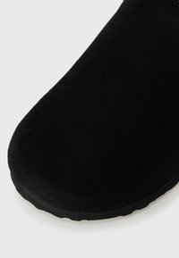 Black suede slipper with a rounded toe and a rubber sole. Smooth texture and minimalist design, featuring no visible hardware or accents.