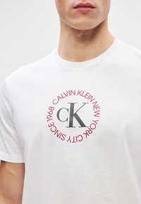 T-shirt de algodão branco com um gráfico redondo vermelho com "CALVIN KLEIN NEW YORK SINCE 1968" e um proeminente logotipo cinza "CK" no centro.