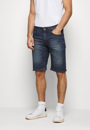Shorts en jean bleu foncé avec une coupe régulière, dotés de cinq poches, d'une braguette à boutons et d'un aspect vieilli pour un contraste de texture. Sneakers blanches aux pieds.