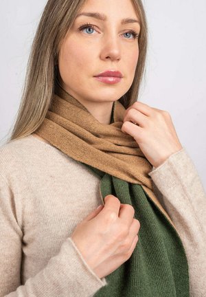 Vrouw in een beige trui die een bicolor gebreide sjaal in camel en groen aanpast voor een effen achtergrond.