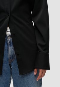 Sort skjorte med knapstil og løs pasform, lavet af glat stof; båret over blå denimjeans. Manchetterne er let oversized, hvilket giver et afslappet udseende.
