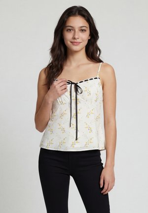 Tânără cu păr lung și închis la culoare, purtând un top alb cu bretele subțiri cu imprimeu floral, cu panglică neagră și pantaloni negri, pe un fundal simplu.