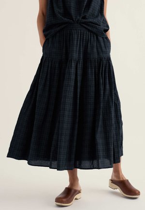 Femeie purtând o fustă midi bleu închis cu volane și buzunare, încălțată cu șlapi maro din piele, stând pe un fundal luminos.