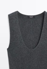 Haut en tricot gris avec un décolleté en V profond, sans manches, et des détails de couture visibles. La texture du tissu semble douce et légèrement extensible.