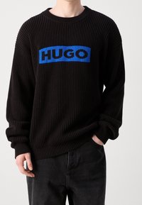Schwarzer Strickpullover mit Rippstruktur, ausgestattet mit einem blauen Grafikband, das "HUGO" in fetten Buchstaben über die Brust zeigt. Bequeme Passform.