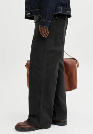 Personne portant un pantalon noir à jambes larges, des chaussures marron, une veste en denim bleu, tenant un grand sac en cuir marron sur un fond uni.