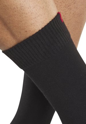 Nahaufnahme von gekreuzten Beinen, die schwarze gerippte Socken mit kleinem rotem Detail am oberen Bündchen tragen.