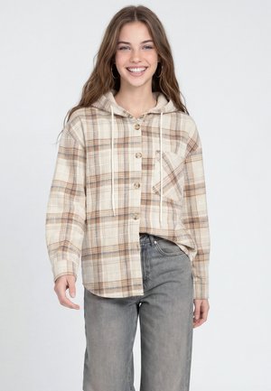Giovane donna sorridente che indossa una felpa con cappuccio beige e marrone a quadri con bottoni e jeans grigi larghi su uno sfondo semplice.