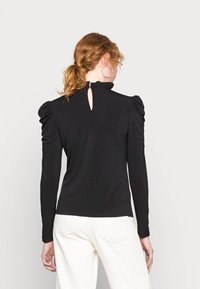 Donna con capelli rossi raccolti in una coda di cavallo, che indossa una maglietta nera a maniche lunghe con spalle a sbuffo e pantaloni bianchi, di spalle su uno sfondo neutro.