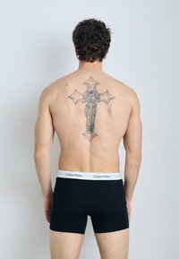 Črne bokser hlačke z belim pasom Calvin Klein na moškem s tetovažo križa na hrbtu. Kratki, kodrasti lasje, obrnjeni stran.