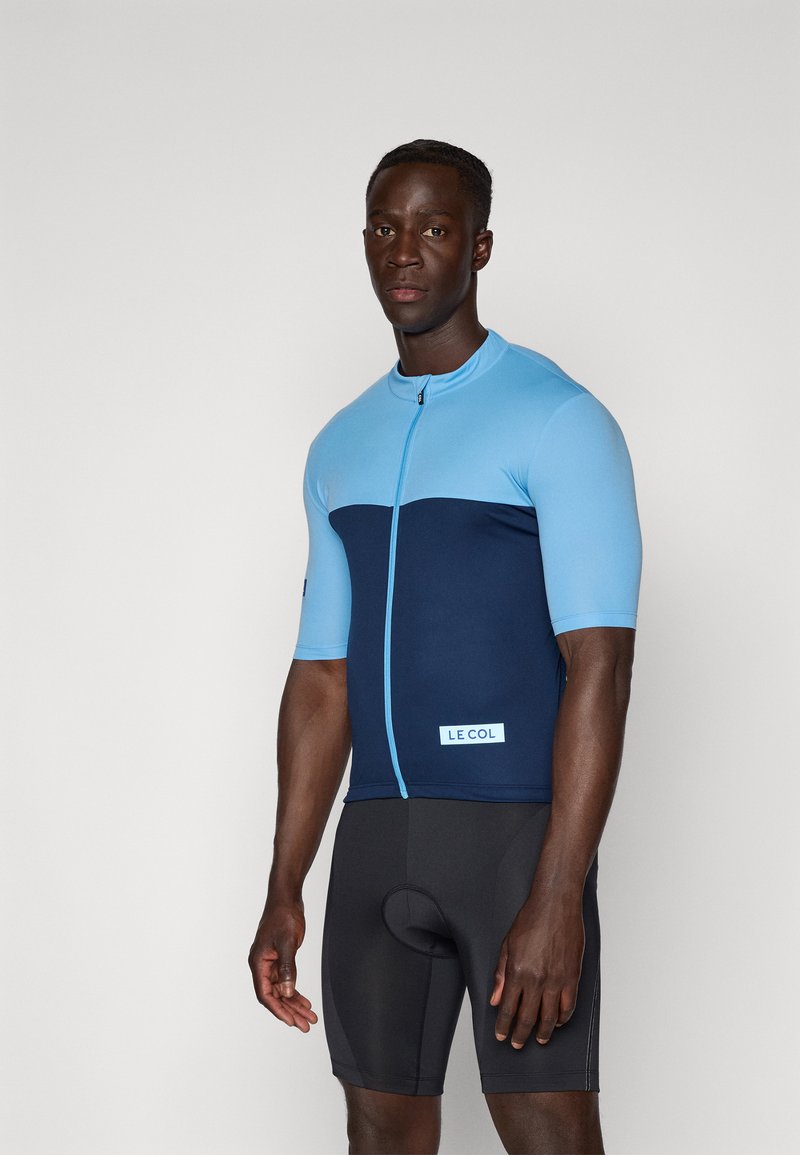 Maillot de cyclisme à manches courtes en bleu clair et marine, avec une fermeture éclair et un tag de logo, associé à un short rembourré noir. Texture lisse.