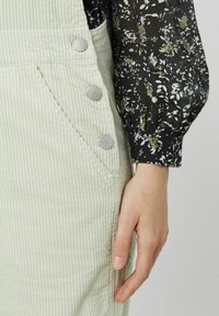 Salopette en velours côtelé vert clair avec des boutons sur le côté, associée à un blouse à motifs floraux sombres avec des manches bouffantes. Détails du tissu texturé visibles.