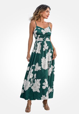 Vrouw draagt een groene maxi-jurk met witte bloemenprint, spaghetti bandjes en doorzichtige sandalen met hak, staand tegen een effen achtergrond.