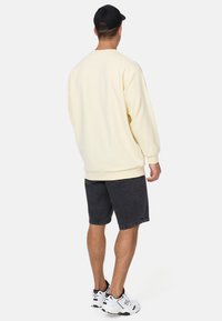 Sudadera de gran tamaño color crema, de material de algodón, con puños y dobladillo acanalados, combinada con pantalones cortos de mezclilla oscura y zapatillas deportivas blancas.