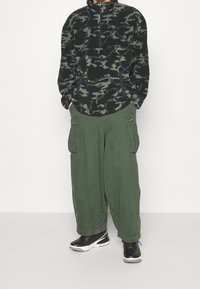 Schwarzer und grüner Camouflage-Fleece-Pullover mit halbhohem Reißverschluss am Kragen, kombiniert mit grünen Cargohosen mit Seitentaschen. Schwarze Sneakers sind zu sehen.