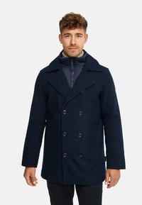 Marineblauer, doppelt geknöpfter Mantel mit breiten Revers, Fronttaschen und großen schwarzen Knöpfen. Darunter eine marineblaue, Zip-Jacke. Textur aus Wollmischung.