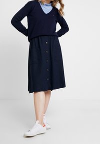 Tmavě modrý svetřík s výstřihem do V v kombinaci s tmavomodrou sukni ke kolenům s předními knoflíky a kapsami. Kombinováno s bílými teniskami s oranžovými doplňky.