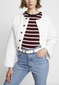 Hvit denimjakke med rund krage og gullknapper, lagd over en svart-og-rød stripete skjorte. Høytlivde blå jeans og et hvitt belte.