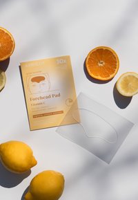 Verpakking van Apricot Vitamin C Forehead Pad, lichtoranje met witte accenten, naast verse citroenen en sinaasappels op een witte achtergrond.