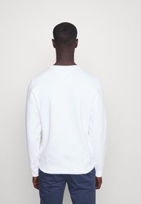 Sweatshirt blanc à col rond, avec des poignets et un ourlet côtelés. Présente une texture unie avec une coupe droite, porté sur un pantalon bleu foncé.