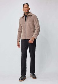 Veste marron clair texturée avec une fermeture éclair à l'avant, un col pointu et deux poches latérales, associée à un pantalon noir et des chaussures avec des semelles marron.