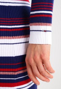 Abito a maglia a coste con righe orizzontali nei colori blu navy, rosso, bianco e dettagli metallici. I polsini sono bianchi con un piccolo tag del marchio.