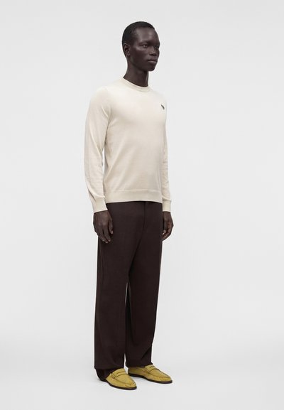 Homme debout portant un pull crème à manches longues, un pantalon large marron foncé et des mocassins jaunes sur un fond clair uni.