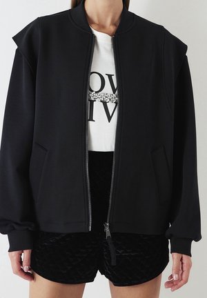 Person iført en sort lynlåstrøje over en hvid grafisk t-shirt med krystaldetalje, parret med teksturerede sorte shorts, stående mod en ensfarvet væg.