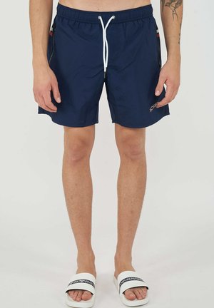 Homme portant un short de bain bleu marine avec un cordon blanc et des sandales blanches Emporio Armani, debout devant un fond uni.