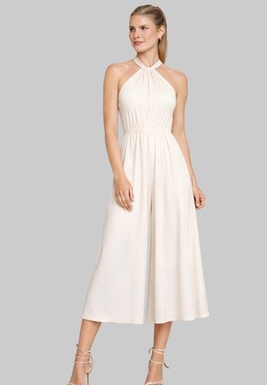 Kobieta ubrana w bezrękawny kremowy kombinezon na szyję z szerokimi nogawkami, stojąca na tle jednolitego szarego tła.