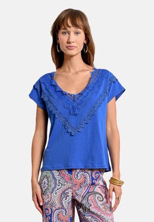 T-shirt in cotone blu con maniche corte, caratterizzato da un bordo in pizzo ondulato attorno a un ampio scollo a V. Abbinato a pantaloni con motivo paisley colorato.