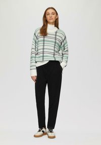 Wit sweater met een groen en zwart ruitpatroon, gecombineerd met zwarte joggingbroek met trekkoord en witte sneakers met zwarte en bruine accenten.