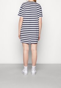 T-paita mekko raidallisena tummansinisenä ja valkoisena. Rennon istuvuuden omaava, lyhythihainen. Yhdistetty valkoisiin lenkkareihin ja nilkkasukkiin, nähtynä takaa.